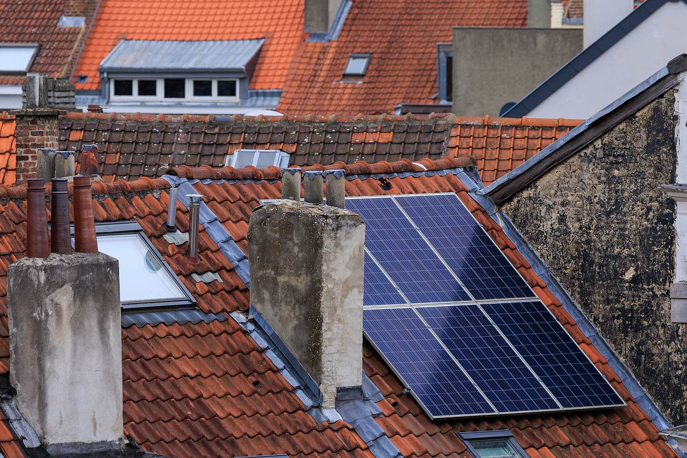 Zonnepanelen op een rood pannendak vlak naast hoge schouwen en een dakvenster, tussen de daken van rijhuizen