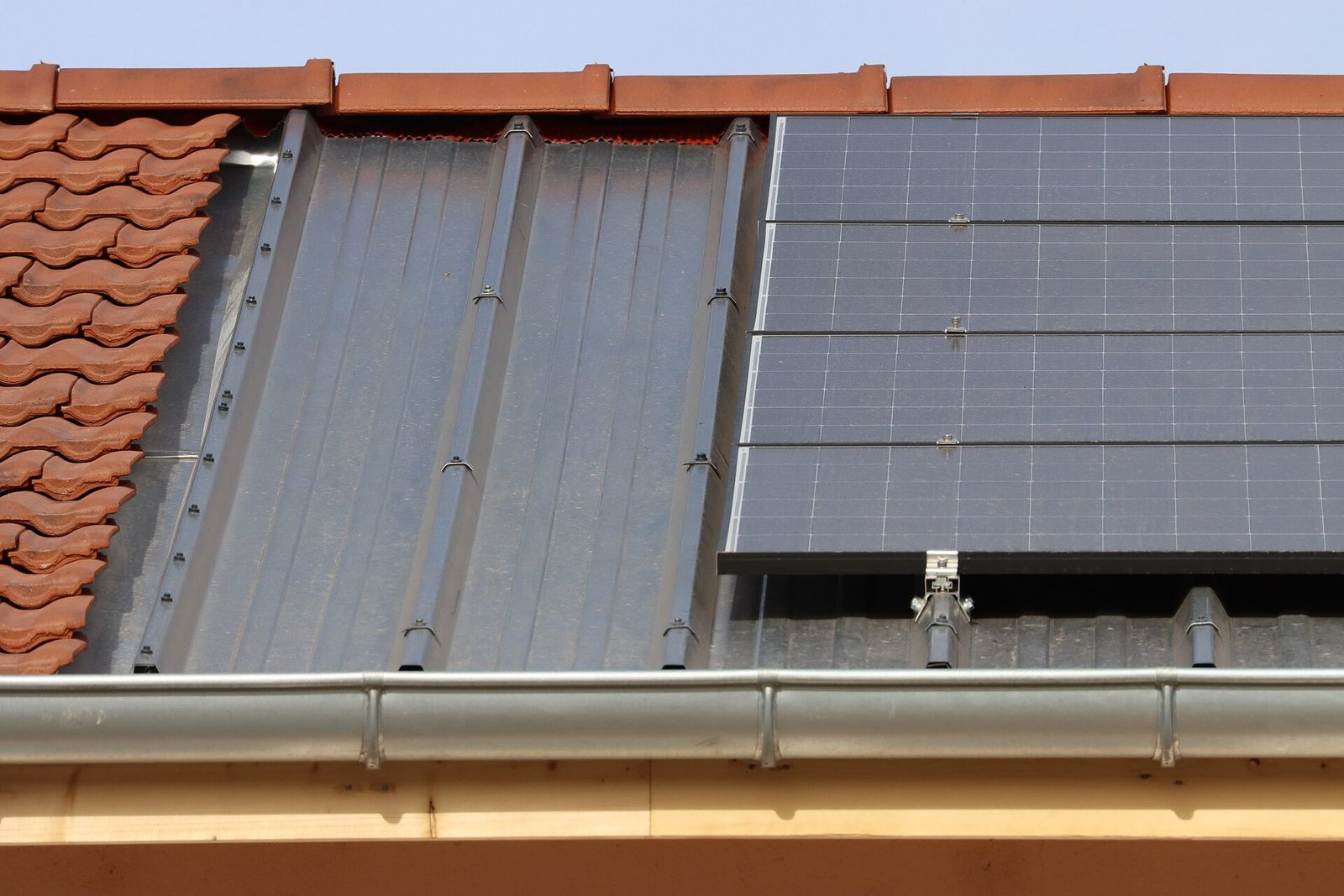 Dakrand waar rode dakpannen overgaan in een metalen onderdak met montagebeugels en een rij zonnepanelen