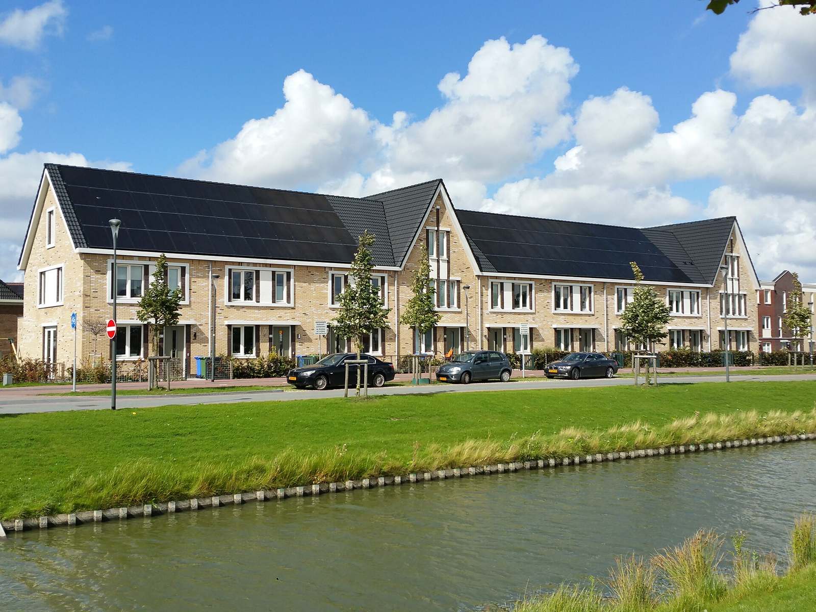 Rij nieuwbouwwoningen met in het dak geïntegreerde zwarte zonnepanelen, gezien over een gracht en een grasberm